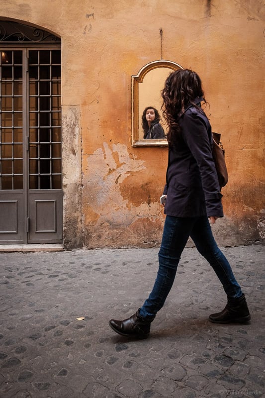 Valérie Jardin photographs a woman walking through the streets in Rome. The girl is looking away from the camera but her face is captured in a mirror facing the camera.