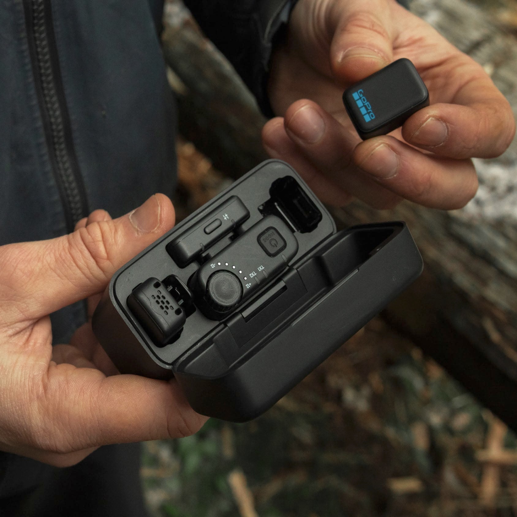 A person outdoors holds an open black electronic device case in one hand and a small rectangular black accessory with a blue indicator in the other hand.