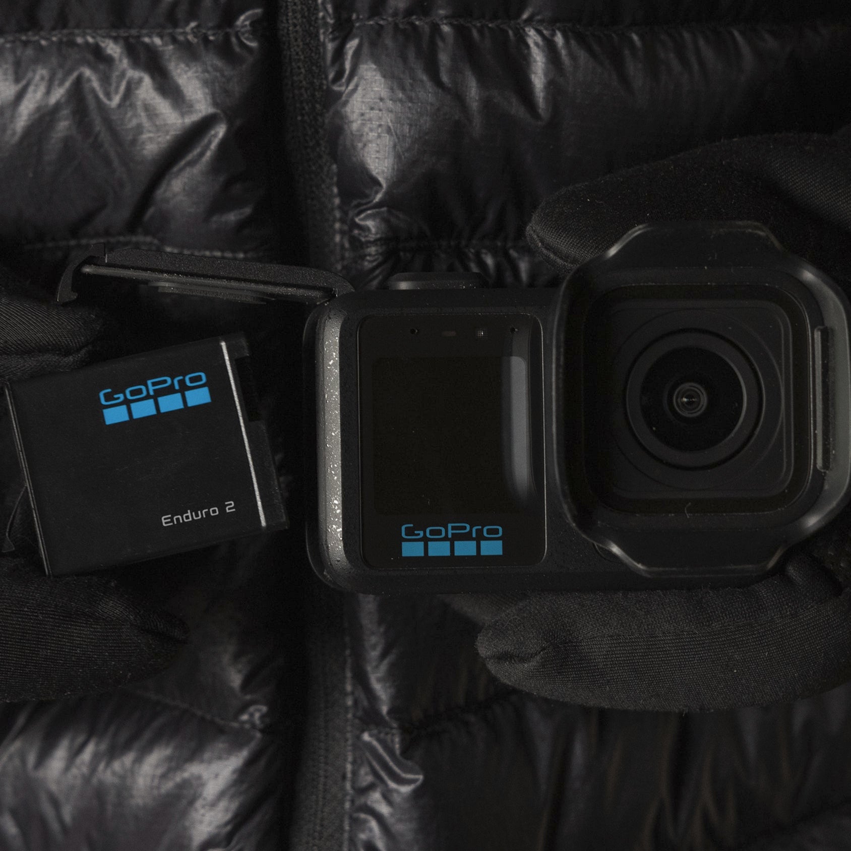 A black GoPro action camera with a black battery pack, both showing the blue GoPro logo, held in gloved hands against a black puffer jacket background.