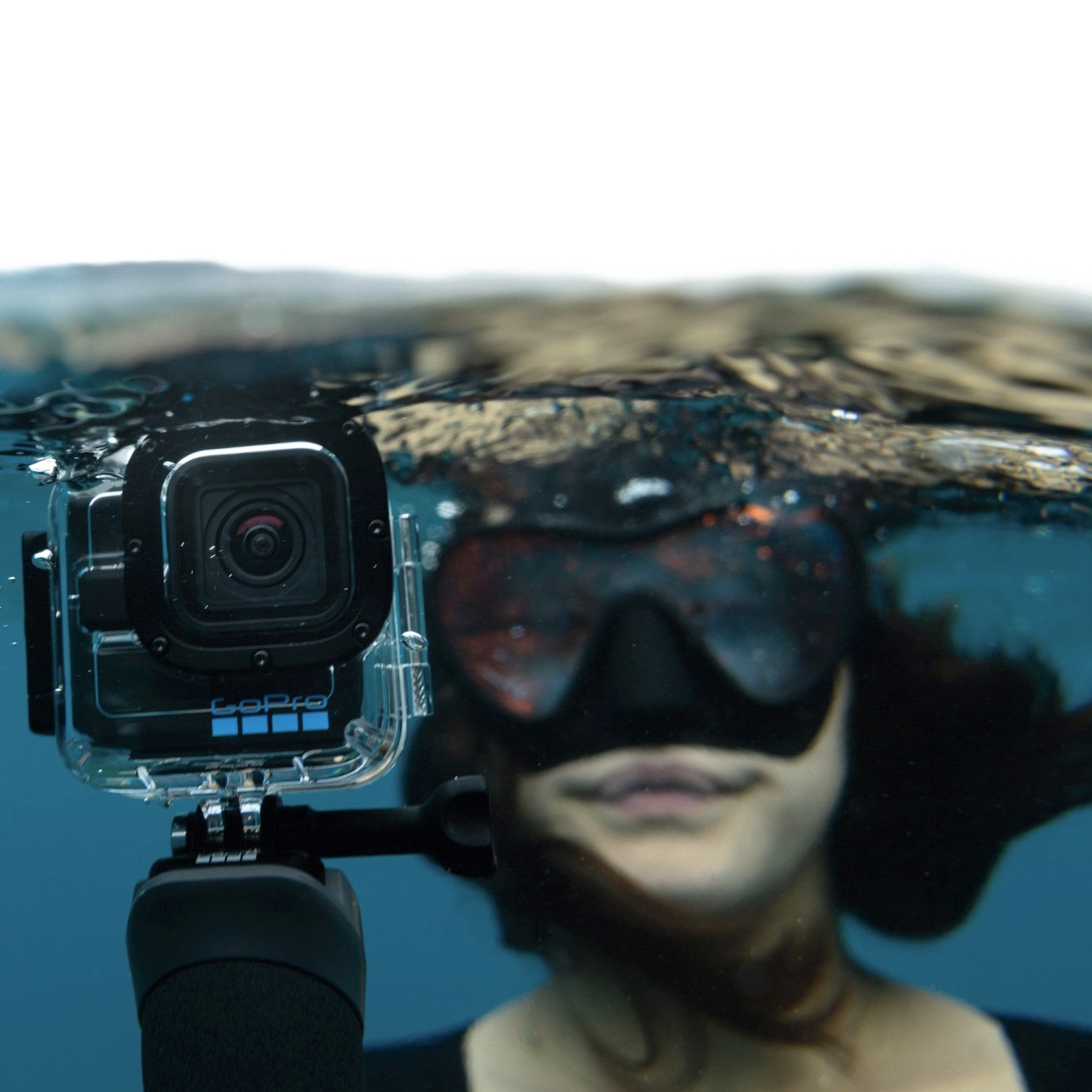 An underwater GoPro action camera in a clear waterproof housing, held by a person wearing a black diving mask and snorkel underwater.