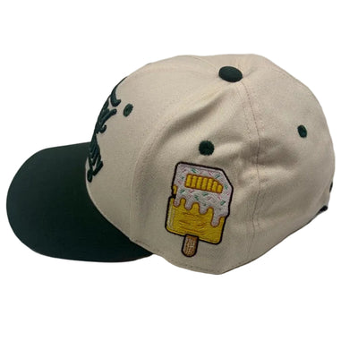 A beige baseball cap with a dark green visor and dark green embroidered details, featuring a yellow and white embroidered popsicle patch on the side.