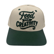 A two-tone baseball cap with a beige crown and a dark green flat brim, featuring the phrase 'Feed YOUR CREATIVITY' embroidered on the front in dark green.