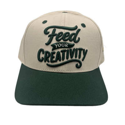 A two-tone baseball cap with a beige crown and a dark green flat brim, featuring the phrase 'Feed YOUR CREATIVITY' embroidered on the front in dark green.