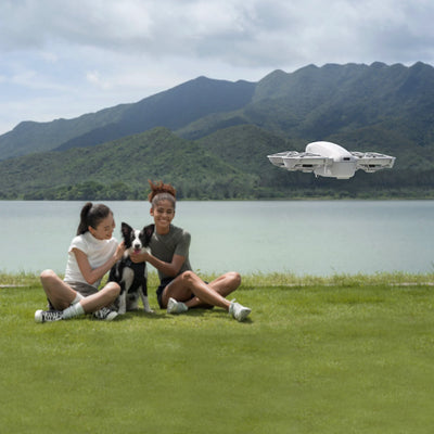 Two people sitting on grass with a dog, a flying drone in the sky, and mountains in the background.